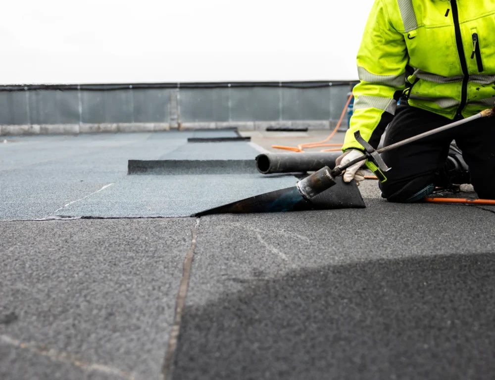 A man in hi-vis gear lays down a strip of flat roofing.