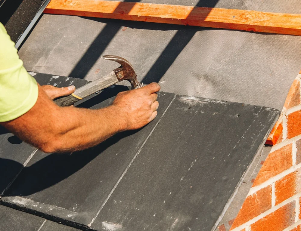 Close up of a man in a green polo shirt hammering a tile into a roofing structure.